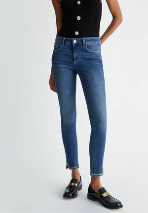 Kvinde iført stramme blå jeans med lynlåse ved anklerne, sort ærmeløs top med knapper og sorte lak-loafers med guldaccent.
