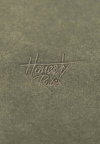 Olivgrön tyg med en texturerad, broderad logotyp som läser "Ärlighet Regler" i en ljus beige tråd.