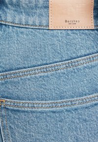 Hellblaue Denim-Jeans besitzen ein braunes Lederetikett mit dem eingeprägten Schriftzug "Bershka". Der Stoff hat eine feine Textur und sichtbare Nähte.