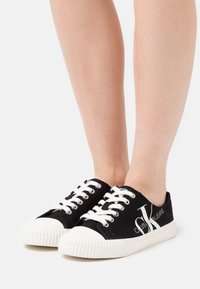 Noha v černo-bílých plátěných teniskách Calvin Klein s bílými tkaničkami a vroubkovanou gumovou podrážkou na neutrálním pozadí.