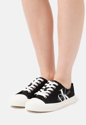 Noha v černo-bílých plátěných teniskách Calvin Klein s bílými tkaničkami a vroubkovanou gumovou podrážkou na neutrálním pozadí.