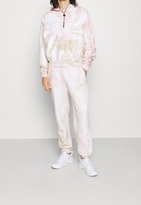 Hoodie in tinta tie-dye rosa chiaro e crema con tasca frontale e cordino regolabile, abbinato a pantaloni jogger coordinati con accenti sui lati.