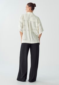 Camisa de corte holgado y manga corta en color crema con patrones de líneas negras, combinada con pantalones anchos de textura negra. Diseño minimalista, sin logotipos visibles.