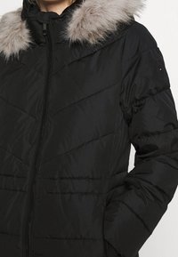 Sort pufferjakke med chevron-syninger, udstyret med en aftagelig faux fur hætte og lynlås-lukning, glat tekstur og sidelommer.