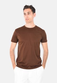 T-shirt marrone a maniche corte realizzato in un tessuto morbido, con scollatura a girocollo e una silhouette aderente. Il modello ha le mani in tasca.