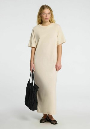 Beige, knee-length T-shirt dress with short sleeves, made from soft fabric. Model holds a black woven tote bag and wears black mesh shoes.