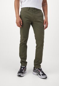 Olive green chinos with a regular fit, smooth texture, and belt loops; worn with black and silver athletic shoes featuring red accents.