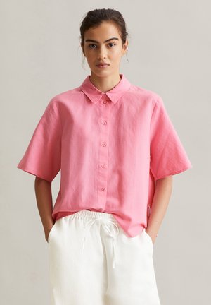 Femme portant une chemise rose à manches courtes et amples, partiellement rentrée dans un pantalon blanc à cordon de serrage, debout avec les mains dans les poches.