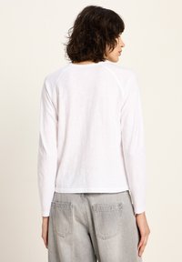 Frau mit kurzen, dunklen, lockigen Haaren, die ein weißes Langarmoberteil und helle graue Jeans trägt, mit dem Rücken zu einem schlichten Hintergrund.