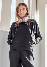 Sudadera negra de manga larga con ajuste holgado y bolsillo delantero, combinada con pantalones deportivos a juego. Auriculares descansan sobre los hombros.