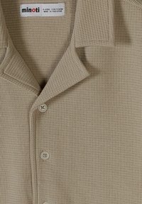 Chemise beige texturée en tricot avec boutons et col, portant une étiquette indiquant minoti, taille 5-6 ans, fabriquée au Pakistan.