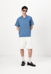 Μπλε κοντομάνικο πουκάμισο από βαμβάκι, λευκές σορτς, μαύρες loafers και κάλτσες. Το πουκάμισο διαθέτει κουμπιά μπροστά και μυτερό γιακά.