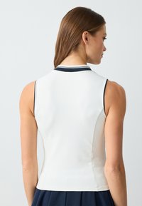 Mouwloze witte polo top met marineblauwe kraagafwerking, getailleerd ontwerp, subtiele textuur en strakke lijnen, met een hoge halslijn en zonder zichtbare logo's.
