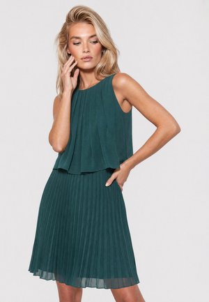 Blonde Frau in ärmellosem türkisfarbenem Plissee-Kleid, eine Hand berührt das Gesicht, die andere auf der Hüfte, mit neutralem Gesichtsausdruck vor hellem Hintergrund.