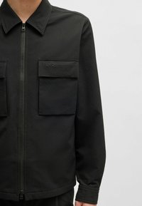 Zwarte zip-up jas met een textuurstof, twee borstzakken en reliëfbranding. Een klassieke kraag en lange mouwen versterken het ontwerp.