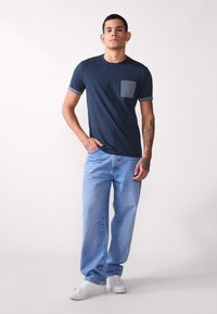 Mornarski T-shirt s uzorkom na džepu, kratki rukavi s usklađenim rubom, uparen s svijetloplavim trapericama i bijelim tenisicama.