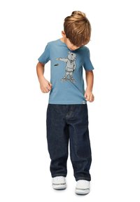 Sininen puuvillapaita, jossa on musta grafiikka karhusta lenkkareissa, yhdistetty tummiin farkkuihin ja valkoisiin kenkiin, joissa on kuvioidut yksityiskohdat.