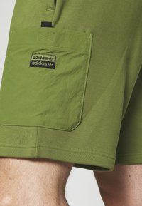 Olivové šortky s velkou kapsou a černým logem Adidas, hladká textura, elastický pás a prošité detaily na okrajích.