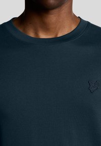 Mørkeblå bomulds t-shirt med rund hals og broderet ørn logo på venstre bryst. Glat tekstur, minimal design.