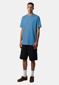 T-shirt blu a maniche corte con un motivo testurizzato, abbinato a pantaloni corti neri e scarpe marroni. La maglietta presenta un dettaglio del logo discreto.
