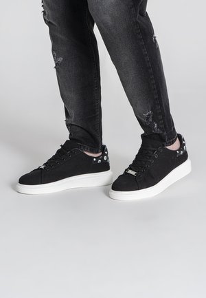 Persona que lleva zapatillas de lona negras con suelas blancas y jeans negros desgastados, de pie sobre una superficie gris lisa.