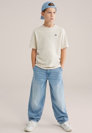 Pamut bézs póló kis fekete logóval, világoskék, bő szabású farmernadrággal párosítva. A megjelenést fehér tornacipő és világoskék sapka egészíti ki.