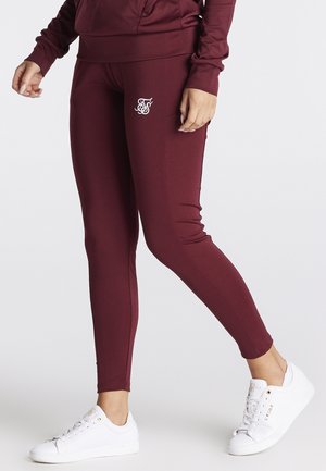 Person iført tætsiddende bordeaux leggings med et hvidt logo på låret og hvide lave sneakers, stående mod en ensfarvet baggrund.
