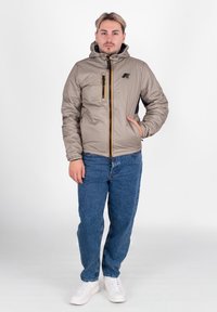 Giubbotto imbottito color beige chiaro con tasca con zip, cappuccio e dettagli neri. Indossato sopra jeans blu e sneakers bianche.