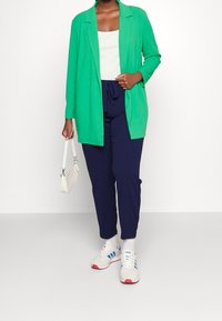 Green long-sleeve blazer over a white top, paired with navy pants. Accessorized with a small white bag and gray sneakers with blue accents.