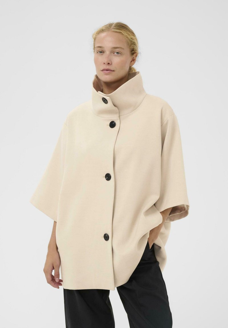 Chaqueta beige de gran tamaño con un collar alto, que cuenta con cuatro botones negros, mangas cortas y una tela suave y texturizada.