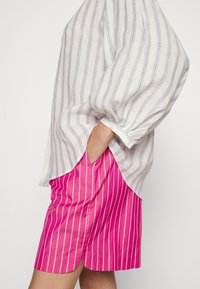 Bílo-modrá pruhovaná lněná košile s volným střihem a manžetami na knoflíky, spárovaná s růžovými pruhovanými šortkami z lehkého materiálu.