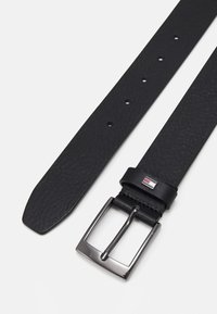 Ceinture en cuir noir avec une boucle métallique rectangulaire et une attache marquée portant un logo. Surface texturée et plusieurs trous de réglage.