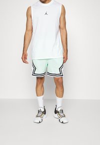 Bílá bezrukávová košile s černým logem, světle zelené síťované šortky s černými a bílými detaily a bílé sportovní boty s barevnými tkaničkami.