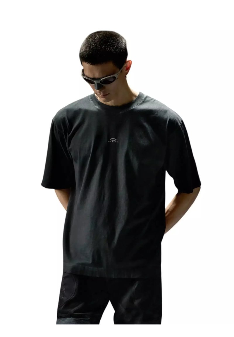 Homme aux cheveux courts portant un t-shirt Oakley noir, un short noir et des lunettes de soleil sportives, regardant vers le bas avec les mains derrière le dos.