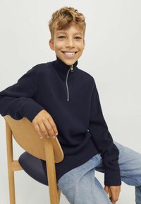 Mornarska zip-up jopica z visokim ovratnikom in rebrasto teksturo, v kombinaciji z svetlo modrimi denim hlačami. Model sedi na leseni stolu.
