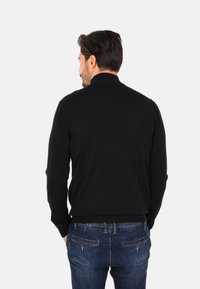 Maglione nero lavorato a maglia con colletto alto, polsini e orlo a coste. Indossato con jeans in denim blu, visto da dietro.