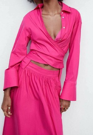 Kobieta ubrana w jaskraworóżowy krótki top w stylu kopertowym z długimi rękawami oraz dopasowaną, marszczoną spódnicę z wysokim stanem, uzupełniona pierścionkami i kolczykami kołami.