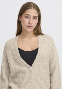 Young woman with long light brown hair wearing a beige textured cardigan over a black top, facing forward with a neutral expression.