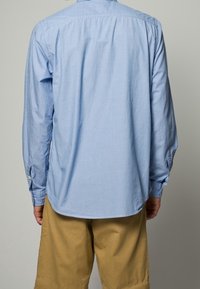 Vaaleansininen raidallinen pitkähihainen nappipaita, jossa on kaareva helma, yhdistetty beigeihin shortseihin. Puuvillamateriaalia, rento istuvuus.