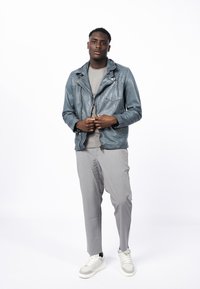 Chaqueta de cuero azul con hombreras y bolsillos con cremallera, combinada con una camiseta gris, pantalones tapered gris claro y zapatillas gris.