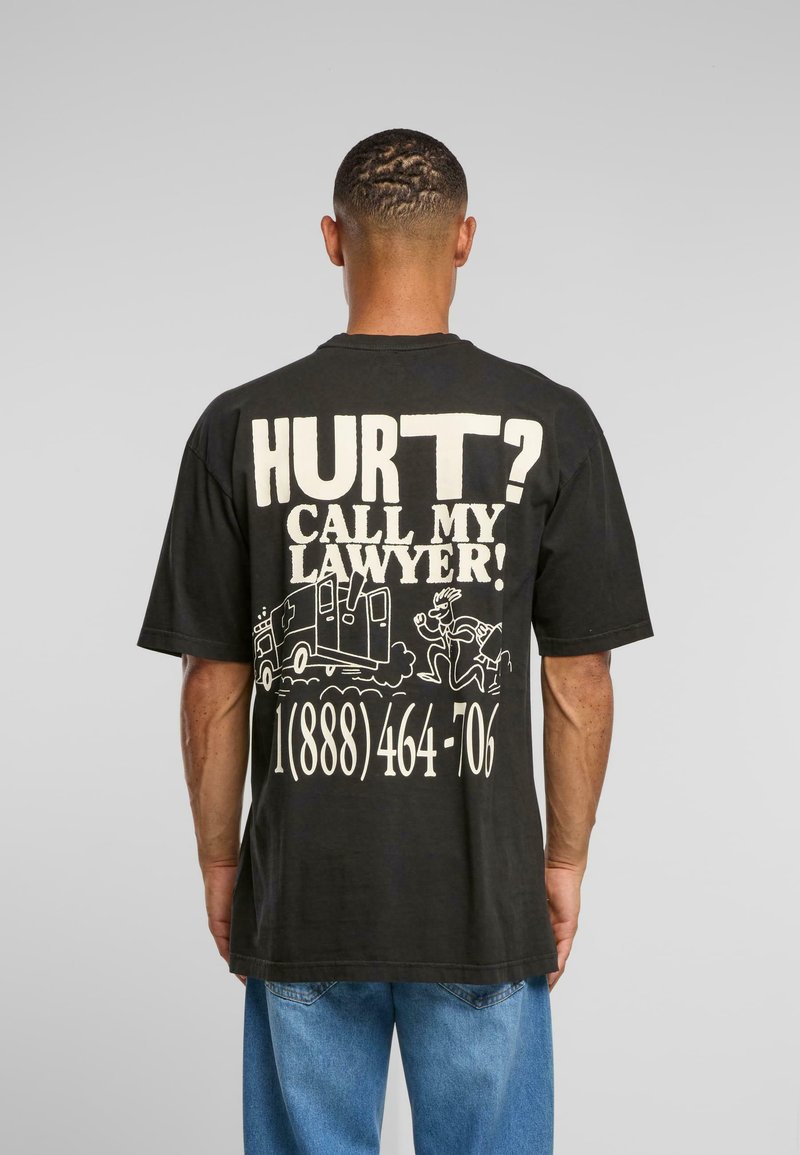 Černé bavlněné tričko s výrazným bílým textem na zadní straně: "BOLÍ? ZAVOLEJ MÉMU PRÁVNÍKOVI!" Obsahuje karikatury a telefonní číslo.