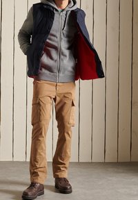 Marineblauw donzen vest over een grijze hoodie, gecombineerd met beige cargobroeken. Bruine veterlaarzen maken de outfit compleet. Achtergrond met houten panelen.