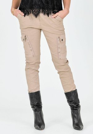 Persoon draagt beige cargobroek met ritszakken, zwarte slungelige leren laarzen en een zwarte kanten top, handen in de zakken gestopt.