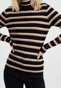 Mujer vistiendo un suéter de cuello alto con canalé y rayas horizontales en negro, beige y blanco, combinado con pantalones negros, con las manos en las caderas.