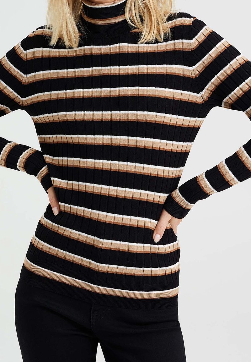 Mujer vistiendo un suéter de cuello alto con canalé y rayas horizontales en negro, beige y blanco, combinado con pantalones negros, con las manos en las caderas.