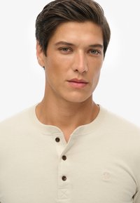 Camisa henley beige con tres botones marrones en el cuello, escote acanalado y logotipo bordado en el pecho. Textura de algodón suave.