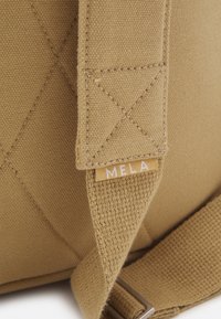 Tan canvas tas met een textuur oppervlak, voorzien van een gestikt patroon en een klein "MELA" label in een contrasterende kleur. Metal hardware zichtbaar.