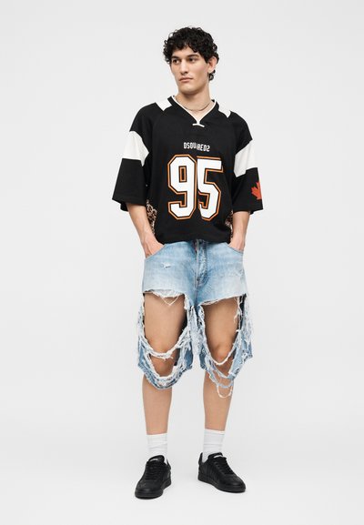 Νέος άνδρας που φοράει μαύρη αθλητική φανέλα με τον αριθμό «95», βαριά σκισμένο ανοιχτό μπλε τζιν σορτς, λευκές κάλτσες και μαύρα αθλητικά παπούτσια, στέκεται με τα χέρια στις τσέπες.