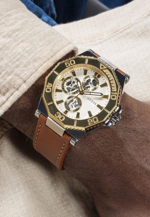 Kullatud ja hõbedane kell, millel on tekstuuriga kreemjas nägu, pruun nahkpael, kaheksanurkne korpus ja kolm silmapaistvat aldekood.