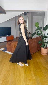 Žena v černých šatech bez rukávů a bílých podpatcích točí se v obývacím pokoji s hnědou pohovkou, velkou zelenou rostlinou a dřevěnou podlahou.